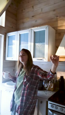 Young woman enjoying her favorite music, dancing in the kitchen in the morning and drinking coffee.