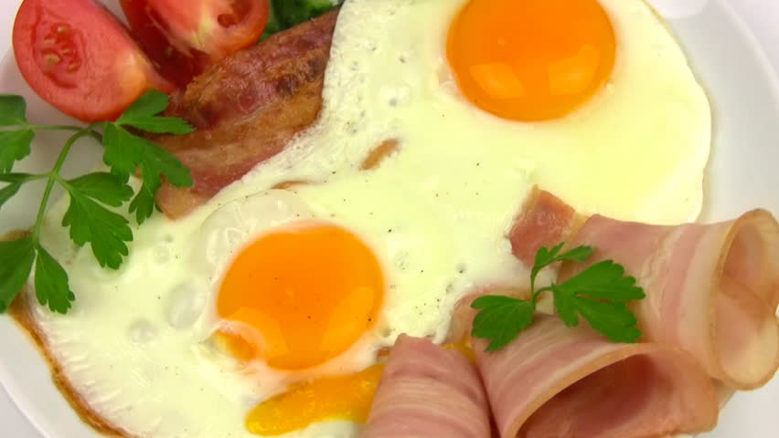 White plate with two fried eggs, bacon, sliced tomato and cucumber, parsley
