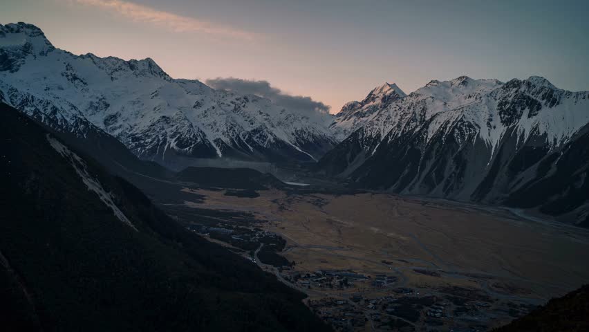 Mountain landscape at dusk or dawn. The mountains are covered in snow, with sharp peaks standing tall against the sky.