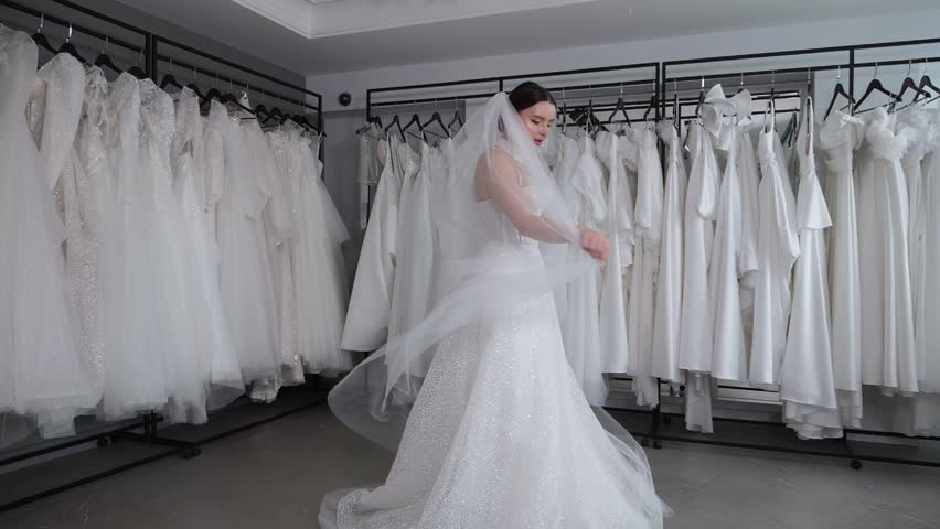A beautiful female model poses in a wedding dress with a long veil in a bridal shop-boudoir, she throws her long veil with her hands. Slow motion.