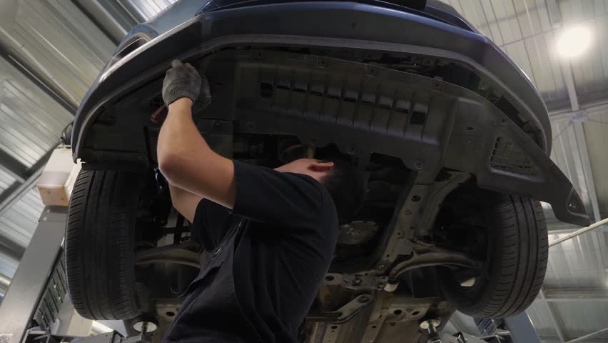 An auto mechanic working under the car on a car lift in the garage, he disassembles the bottom of the car for further repair. Antifreeze drain in the service station. Auto repair shop, auto repair.