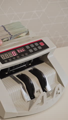 A counting machine rapidly processes stacks of cash in a modern financial office. The digital display shows the number of bills counted. Vertical video.