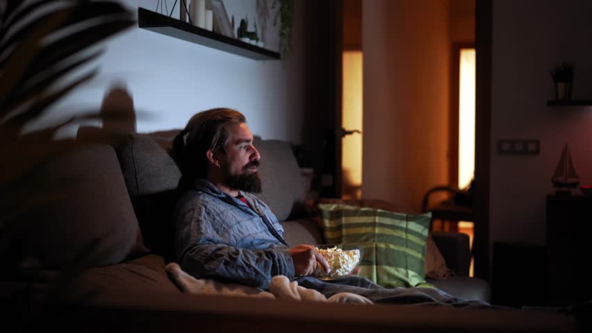 Side view footage of a man sitting on the sofa at home watching movies and eating popcorns. Copy space video.