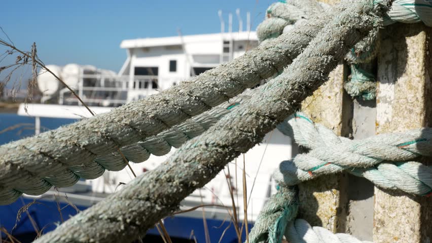 Weathered, moss-covered marine ropes wrapped tightly around a concrete posts. Pier. Fence. Dry grass contrasts with the sea elements and blue sky.