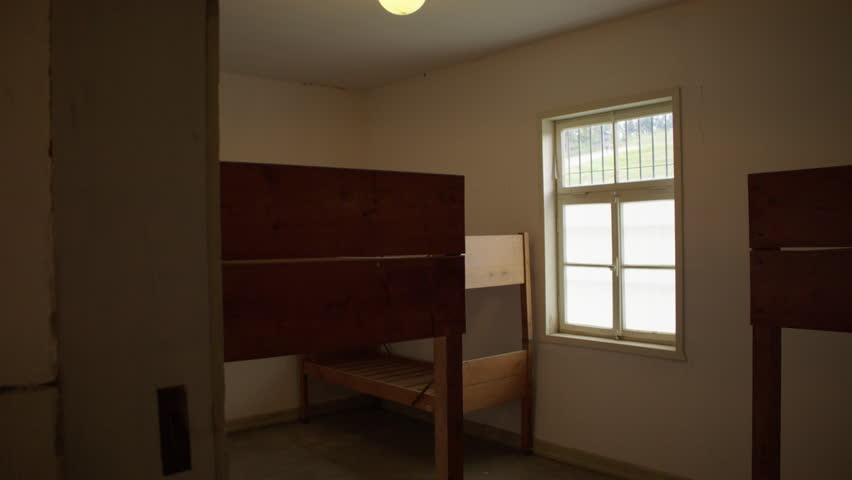 A small room with wooden bunk beds, a concrete floor, and a barred window. The room is sparsely furnished and has an austere atmosphere.
