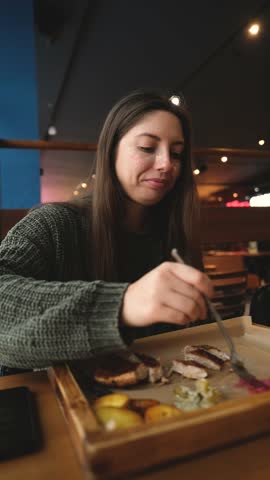 Pretty brunette enjoying tasty beef steak in restaurant in winter. Woman in warm cozy sweater eating meat near window. Snow outside