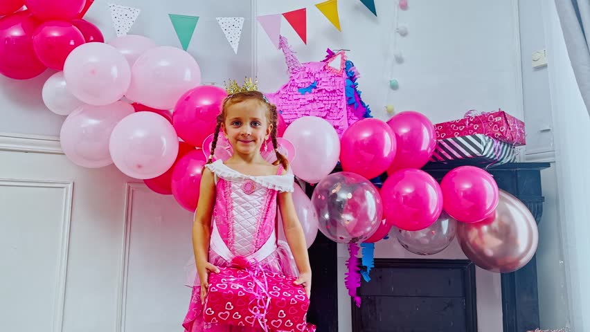 Smiling in her pink princess costume, the girl holds a heart-decorated present, show it to camera surrounded by a cheerful array of colorful balloons