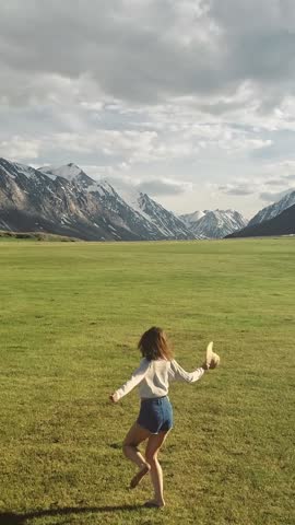 Vertical video. Happy young girl is skipping across the field. Mountains and sky are in the background. Cheerful young woman running with hands stretched wide, breathing deeply, feeling happy being in