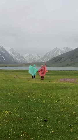 Vertical video. Couple of tourists holding hands standing in grass in rain on background of lake in mountains. Young man and woman in casual clothes and raincoats standing