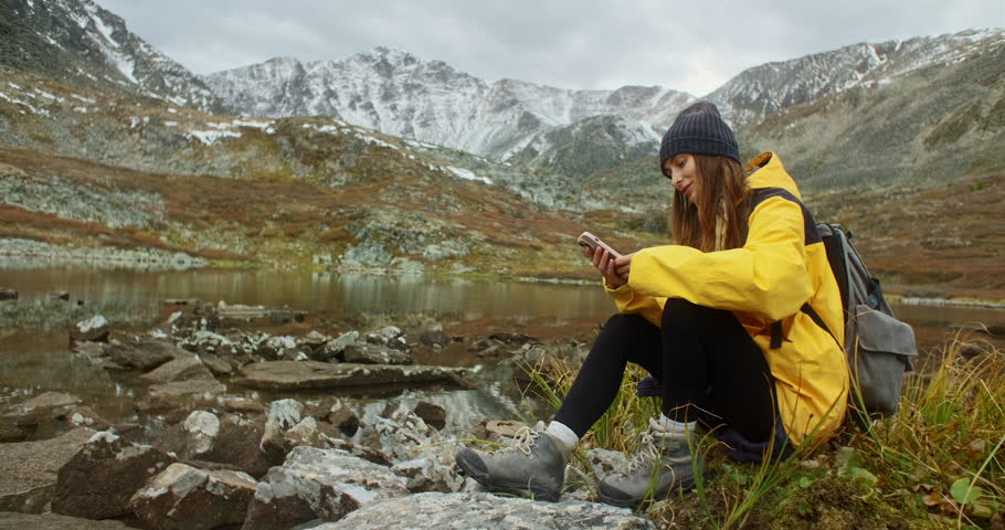 Young backpacker woman sitting by a mountain lake, using her smartphone while soaking in the breathtaking landscape on a cloudy autumn day filled with tranquil beauty
