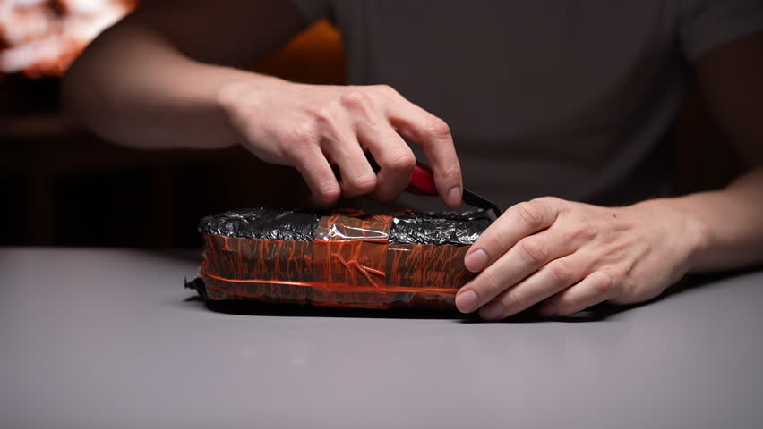 Cropped shot of man carefully opening package containing new mobile phone, using cutter to remove protective plastic and bubble wrap, revealing gadget inside sitting at table, close-up, slow motion.
