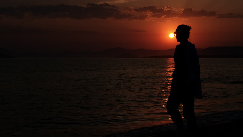 Woman walking towards setting sun on beach. Silhouette of a woman walking along a beach, embracing the beauty of a vibrant sunset. The sky glows with hues of orange, red, and yellow