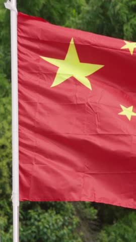 Vertical Slow motion of a Chinese flag waves against a lush green background of trees, vibrant red color and golden stars. Natural setting contrasts beautifully with bold colors of national flag