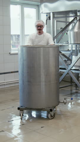 Full vertical shot of Caucasian male cheese factory employee in white apron and cap transporting milk curdling tank on wheels around processing facility