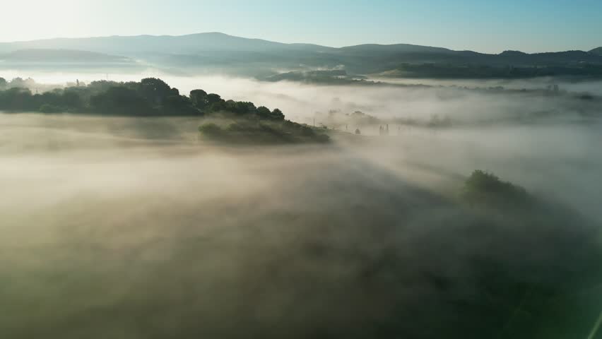 Aerial drone footage of a serene Tuscany sunrise, with golden rays piercing the mist over lush hills and treetops, creating a dreamlike spring landscape.