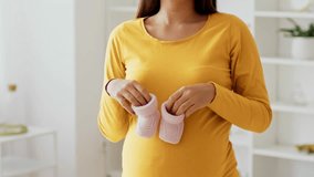 An expectant mother wears a yellow long-sleeve shirt, gently cradling her baby bump while holding a pair of small pink baby shoes, radiating happiness and anticipation in her home. - Powered by Shutterstock - Get 15% off with code: PIKWIZARD15