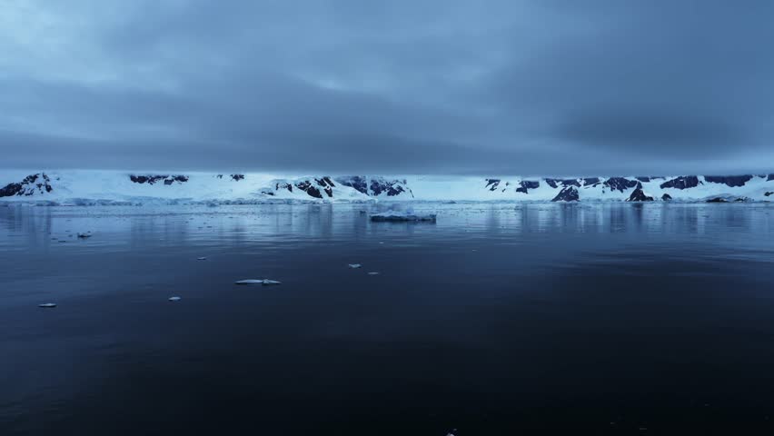 Aerial Drone Shot of Antarctica of Mountains and Glacier with Dramatic Blue Clouds, Antarctica Peninsula Beautiful Scenery on the Southern Ocean Sea with Calm Still Flat Water with Reflections