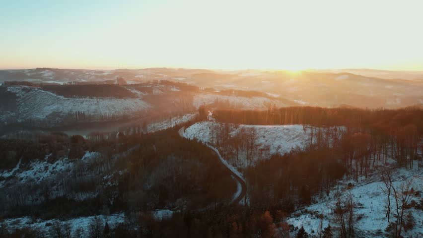 A drone flies over a quiet lake and snowy forest, with golden hour light adding warm tones to the winter landscape.