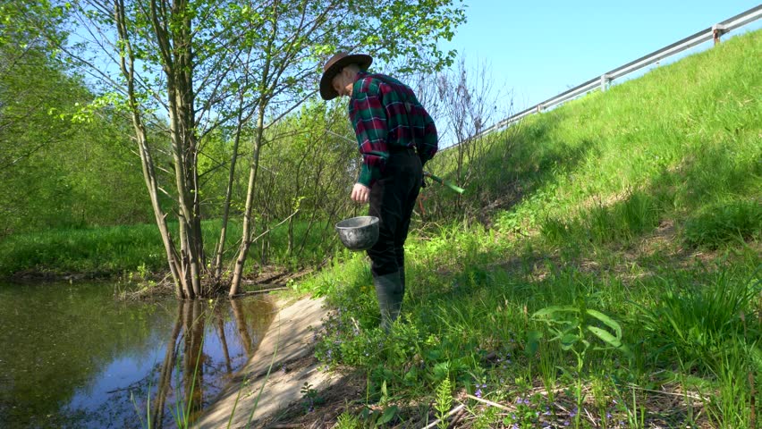 A fisherman casts a baited line into a lake, wearing a cowboy hat, a plaid shirt, grey trousers and rubber boots. It is a sunny day outside and there are lots of insects flying around.