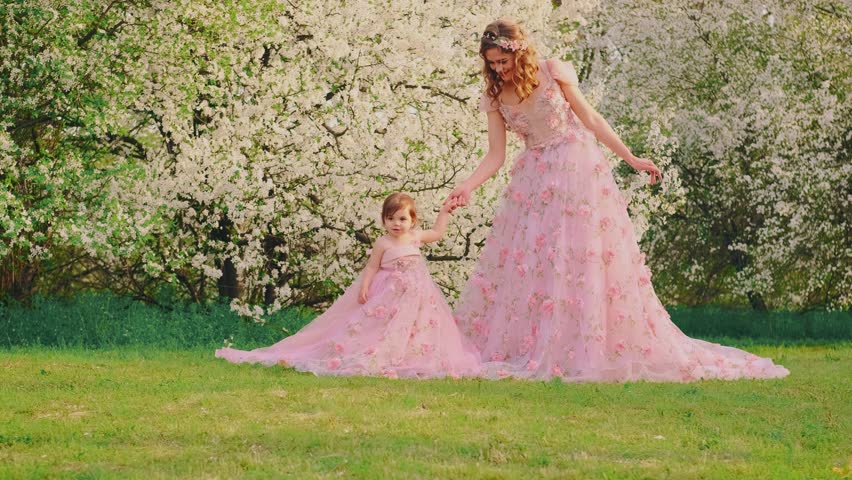 family posing for a photo shoot smiling face woman holding hand little girl. Happy mom and daughter together. Long pink dress. queen with baby princess. spring cherry trees white flowers spring nature