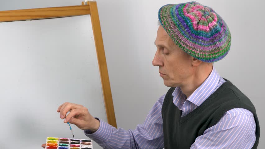 An elderly artist in a striped blue shirt, a gray sweater and a knitted colorful beret applies the first brushstroke to a painting.