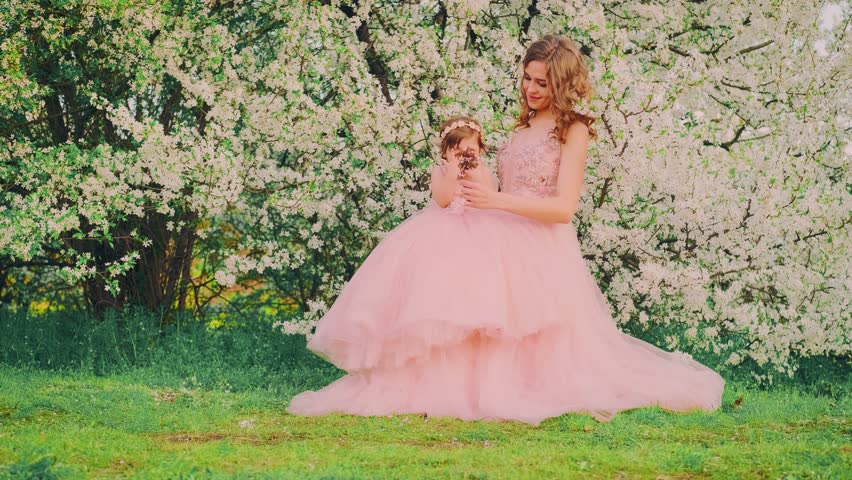 family posing for a photo shoot smiling face woman holding hand little girl. Happy mom and daughter together. Long pink dress. queen with baby princess. spring cherry trees white flowers spring nature