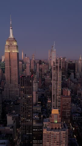 Vertical aerial views capture the Empire State Building in Midtown Manhattan lighting up New York City skyline at dusk, as the sun sets over the lively metropolis