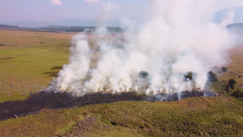 Smoke billows from a controlled fire burning through grassy fields in a vast open landscape