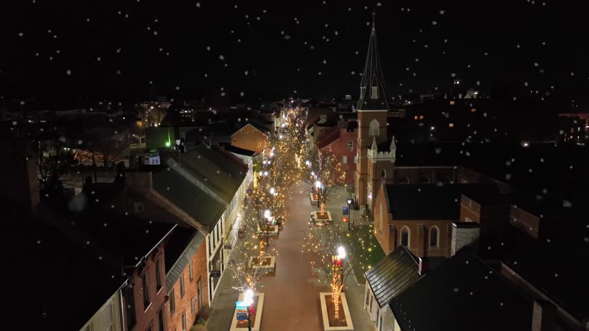 Christmas shopping in small historic town in USA. Lights and snow at night. Snowstorm during holiday season. Historic church and lighting christmas trees. Rising drone wide shot.