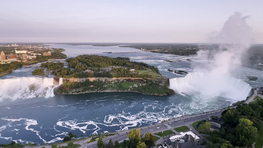 Stunning wide aerial view of American and Canadian Niagara Falls at sunset, with cascading waterfalls, the vibrant river, and a warm pink and purple sky, Canada, zoom out