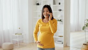 Asian pregnant woman wearing a yellow long-sleeve shirt stands in a well-lit living room, gently cradling her belly while engaging in a phone conversation, creating a warm and joyful atmosphere. - Powered by Shutterstock - Get 15% off with code: PIKWIZARD15