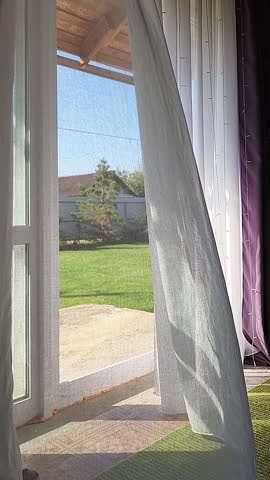 Room interior with open window overlooking back yard with lawn. Transparent tulle with morning sun rays on shadow background. Vertical video. High quality footage