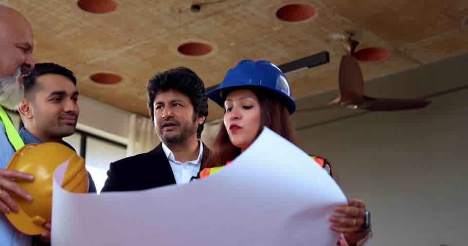 Indian Asian group of civil engineers, architects in fluorescent jackets, hard hats, and corporate investors discussing project plans together over blueprints in a modern corporate office setting
