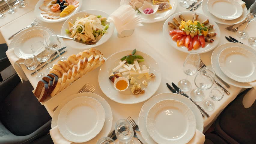 Table with a variety of food and drinks, including bread, cheese, and wine glasses. The table is set for a meal, and there are several forks and knives on the table. Scene is inviting and welcoming