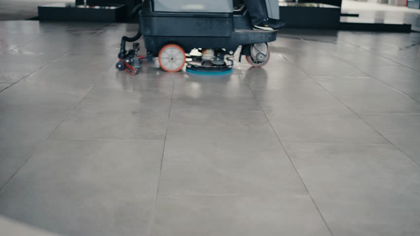 A person uses a floor scrubber to clean a tiled floor. The machine leaves the floor shiny and clean. Perfect for cleaning services, hygiene, and maintenance projects.