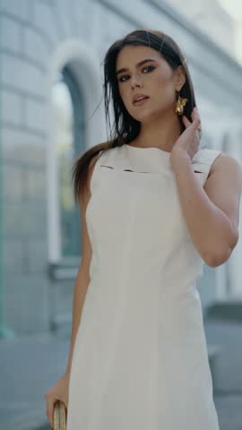 Young, beautiful woman in white dress poses on city street. She holds her hand by face and looks around. Slow motion shot of elegant lady enjoying sunny day outdoors.