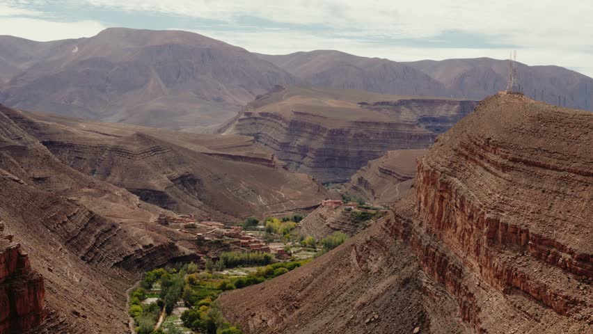 Breathtaking aerial view of a small village nestled in the Dades Gorge, Morocco. The lush greenery contrasts with the rugged, layered rock formations of the High Atlas Mountains,