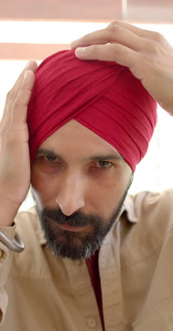 Vertical video: Adjusting red turban, man looking at camera with serious expression. Portrait, tradition, culture, headwear, ethnic, fashion