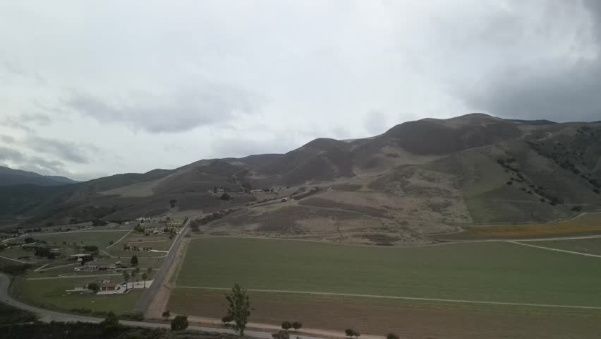 Aerial view capturing the Arroyo Seco river winding through the agricultural landscape of Greenfield, California, under a cloudy sky and gentle hills, drone pannig