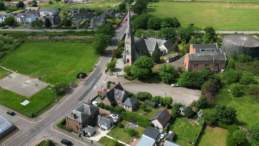 Drone footage of Invergordon showcasing a church with a tall spire, surrounded by green spaces, residential homes, and industrial tanks in the scenic Scottish Highlands.