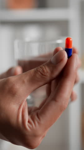 A hand grasps a colorful capsule poised above a clear glass of water. The setting suggests an indoor space, focusing on health and wellness, emphasizing self-care