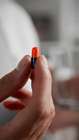 A person holds a bright orange and blue capsule in one hand while grasping a clear glass of water in the other hand. The setting appears to be a home or healthcare environment