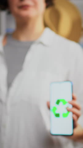 A person stands in a well-lit indoor space, proudly displaying a smartphone featuring a green recycling symbol on its screen. The focus is on promoting sustainability