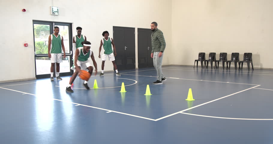 Multiracial male basketball players bouncing basketball and avoiding cones with coach in gym. sport, lifestyle and activity.