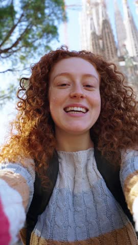 Vertical. Young caucasian redhead women taking a selfie in front of Sagrada Familia in Barcelona. Smiling beautiful girl on street of European city enjoying vacation happy and smiling. Tourist people