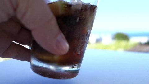 a hand takes a glass and pours cola in a glass filled with ice cubes and some piece of a lemon