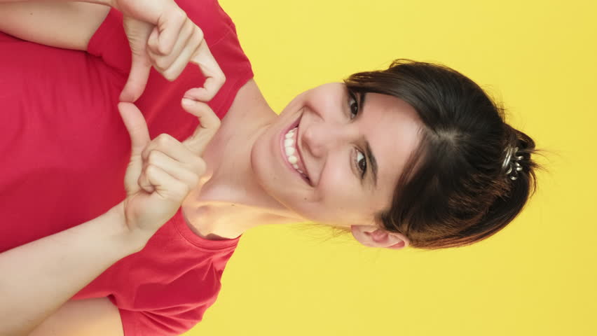 Vertical video. Heart symbol. Air kiss. Woman showing love heartfelt adore amour hand gestures affectionate body language isolated on yellow free space background.
