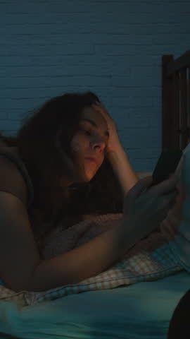 Vertical video. Insomnia and sleep problems. Person with smartphone in her hands lying awake on bed at night.