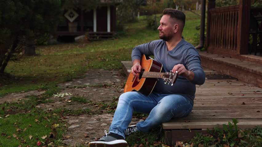 50 years old man with dark hair in a gray shirt sitting on the wood terrace while playing guitar, musical instrument in park. Male guitarist in blue jeans resting and enjoying sunset light