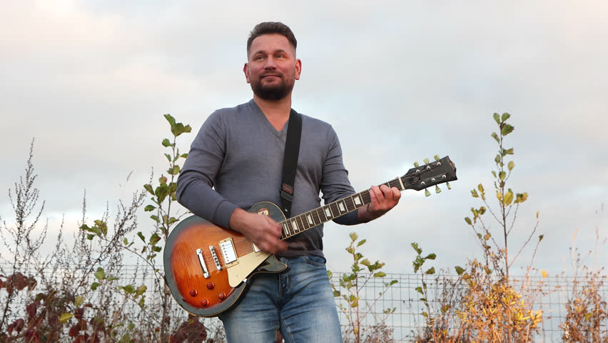 50 years old man with dark hair in a gray shirt standing on the grass having joy while playing bass guitar, musical instrument in park. Male guitarist in blue jeans resting and enjoying calm atmospher